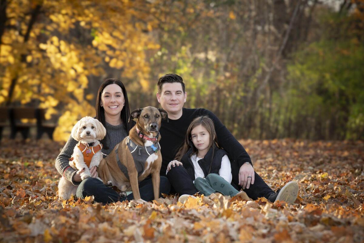 a family of three sitting outdoor on yellow springtime leaves with their two dogs