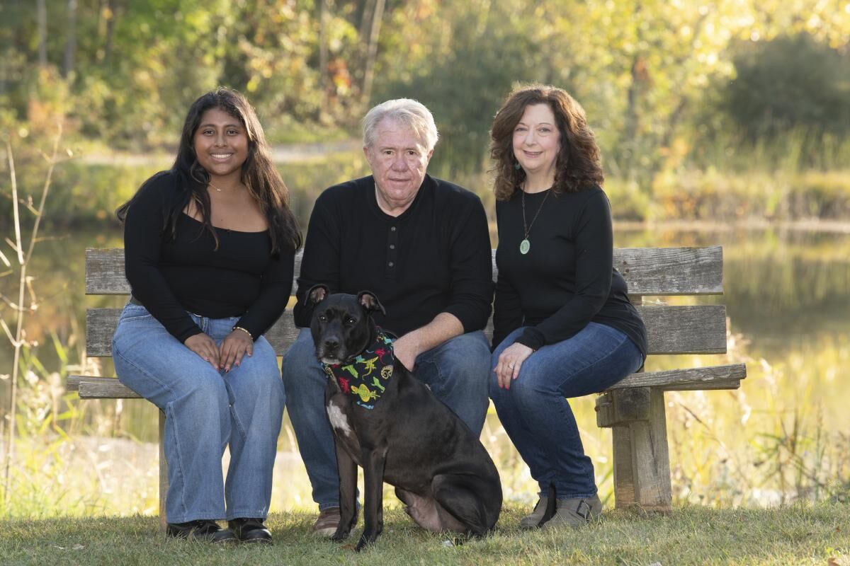 A middle-aged woman with long dark hair, an elderly man with short gray hair, and a young woman with long dark hair sitting on a bench outdoors with a black dog