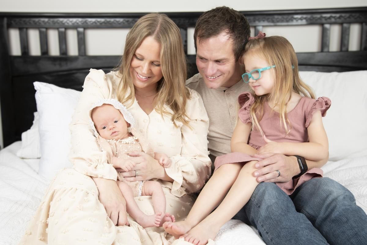 A young family of four - a mother, father, and two young daughters, one a newborn baby and the other a toddler, sitting together on a couch and smiling