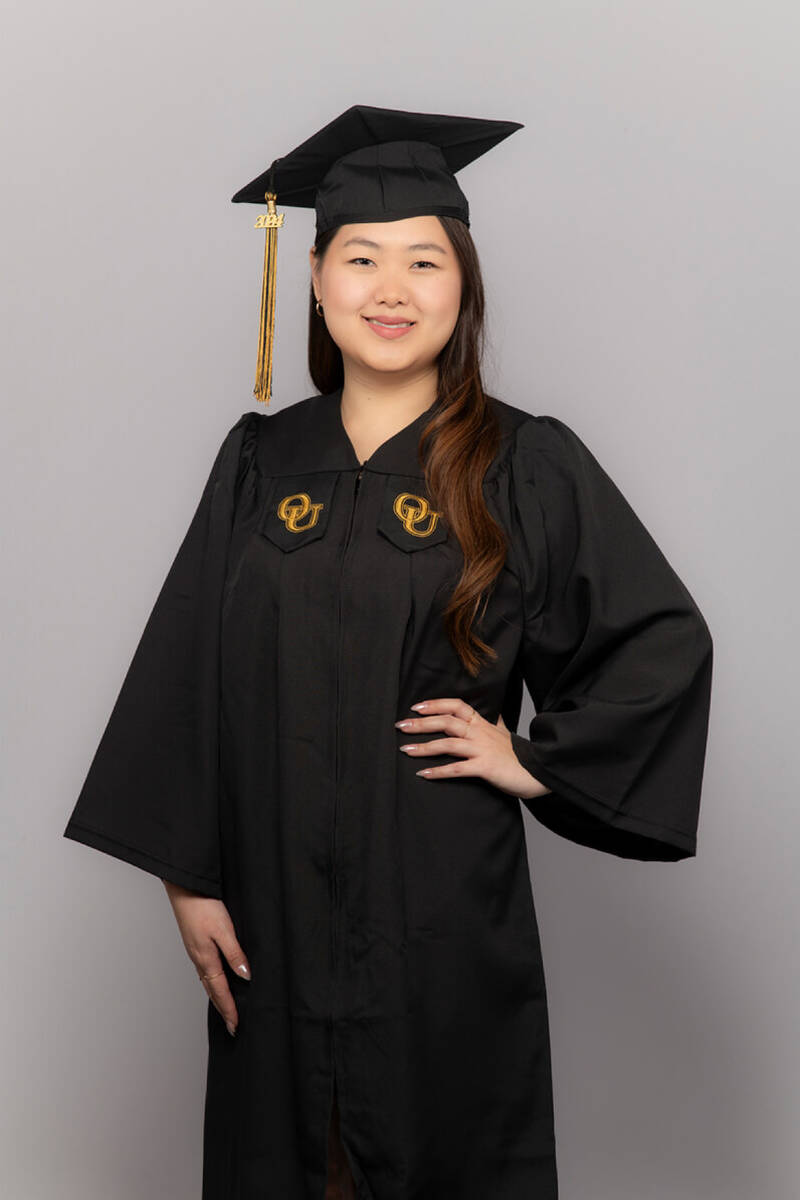 A brown-eyed, dark-haired girl in a black graduation gown with a square master's cap. Behind her is a gray background